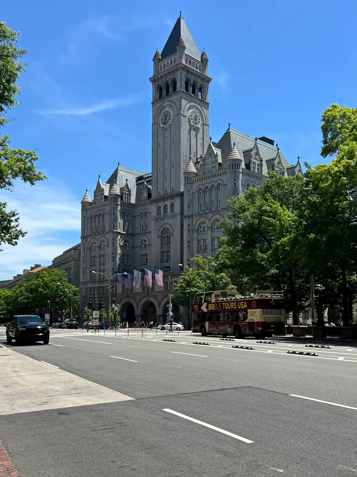 The Old Post Office Pavilion’s gray stone clock tower rises above the street, with steep slate roofs, arched windows, and small turrets framed by leafy green trees under a bright blue sky. Flags hang over the entrance while a double-decker tour bus and cars pass in the foreground, giving the scene a lively downtown feel.