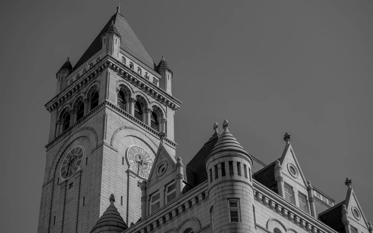 Low-angle black-and-white view of the Old Post Office Tower, with its steep slate roof, arched belfry openings, and twin clock faces rising above the ornate stone facade. The dramatic composition isolates the historic architecture against a blank sky.