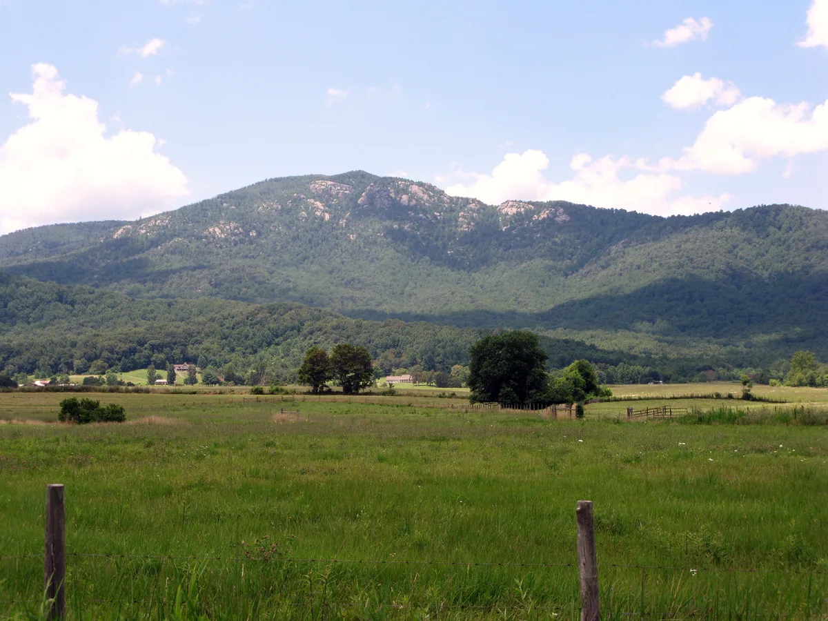 Sunlit green fields stretch across the foreground beneath Old Rag Mountain’s rounded, rocky summit and forested Blue Ridge slopes. Puffy white clouds drift across a pale blue sky, with scattered farm buildings and fence posts adding scale to the wide valley view.