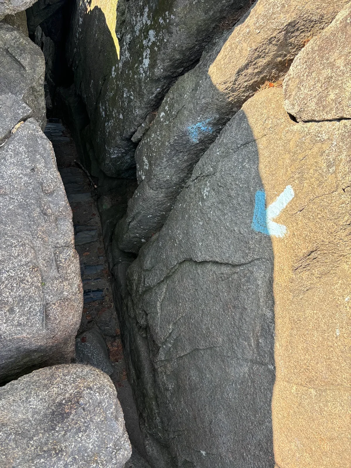 A narrow rocky chute at Old Rag Mountain, with massive gray granite boulders squeezing a steep passageway in deep shadow. Sunlight warms the right-hand rock face, and blue trail blazes mark the route through the crevice.