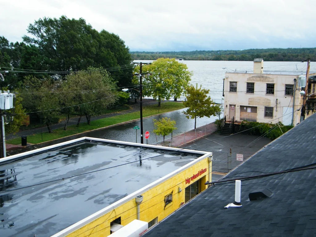 Looking down from an upper floor over a rain-slicked waterfront street, the photo shows a yellow building with a flat roof in the foreground, a row of trees and a narrow road leading to the Potomac, and a pale historic building at the water’s edge. The overcast light softens the river and distant shoreline beyond.