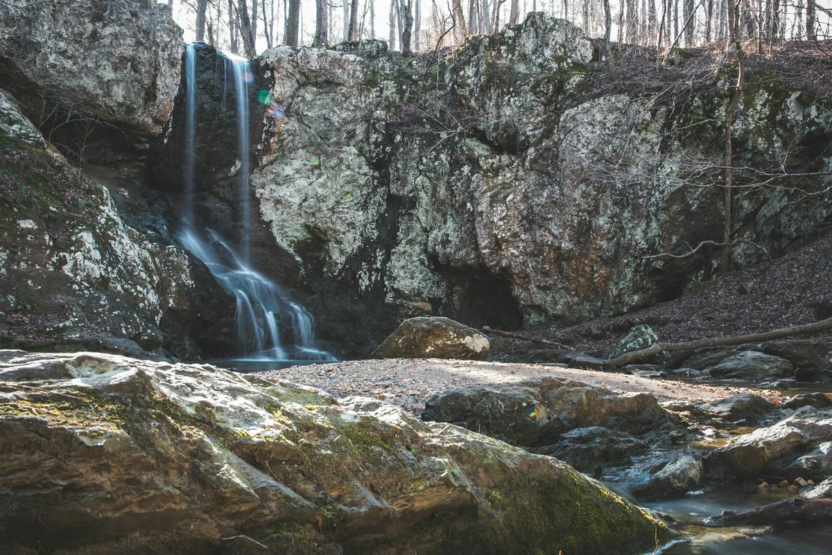 A narrow, long-exposure waterfall drops over a dark rock ledge into a shallow pool, framed by mossy boulders and a steep, lichen-speckled cliff. Bare winter trees rise above the gorge, and sunlight catches the foreground rocks and trickling stream below.