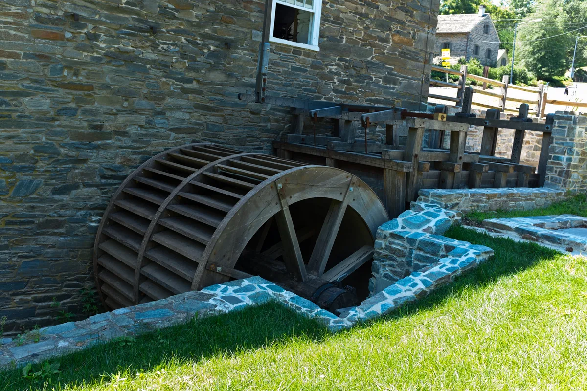 A restored wooden waterwheel sits beside the rough stone wall of Peirce Mill, partly tucked into a shaded millrace lined with pale blue-gray masonry. Bright green grass and sunlit stone steps frame the old mechanism, with a small stone outbuilding and trees in the background.