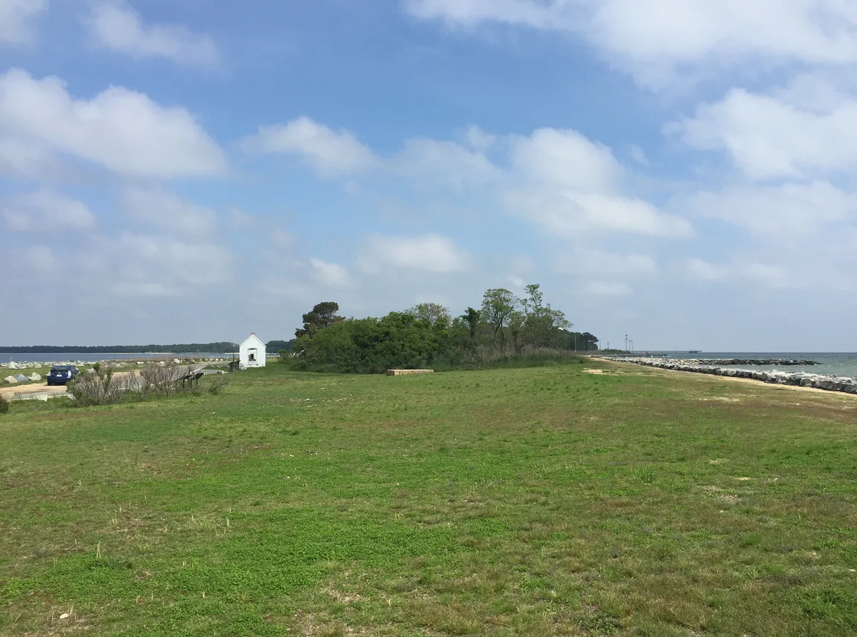 Wide view from Point Lookout Lighthouse across a grassy peninsula to the Chesapeake, with a small white outbuilding and low trees near the center. Rock-lined shoreline borders the water on both sides under a bright blue sky filled with scattered clouds.