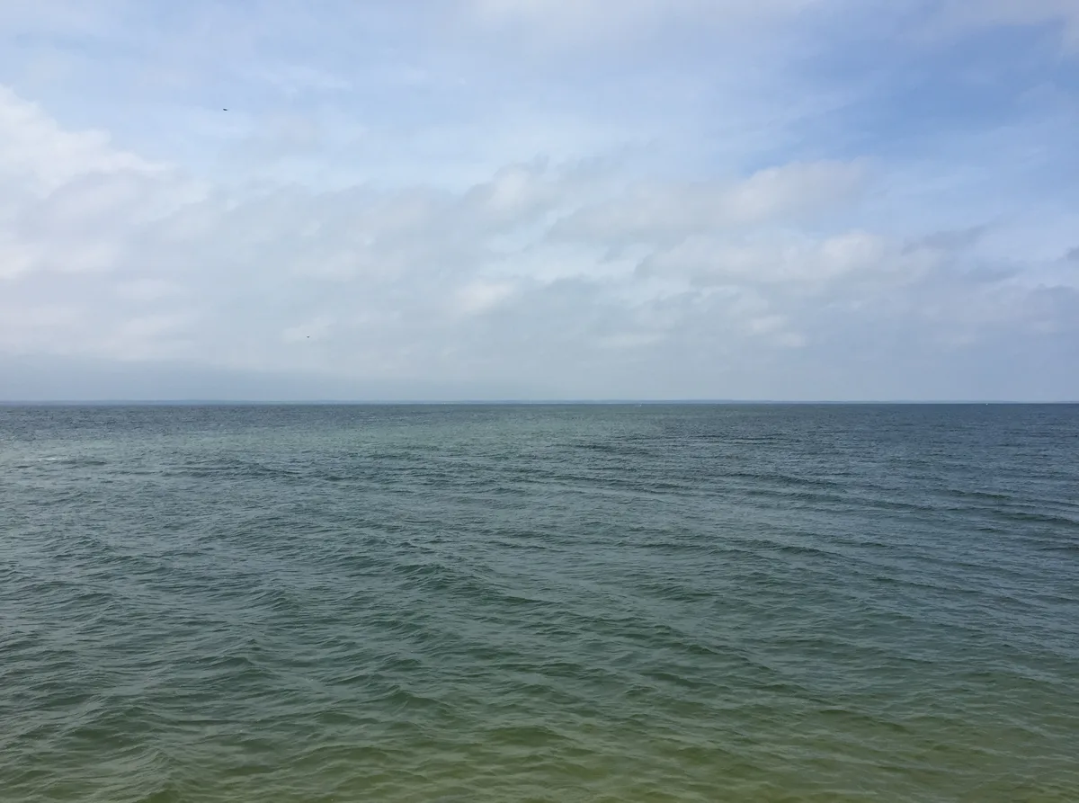 Wide view over choppy blue-green water under a pale, cloud-streaked sky, with a thin, distant horizon stretching across the frame. The open scene feels expansive and calm, with no land or lighthouse visible in the shot.