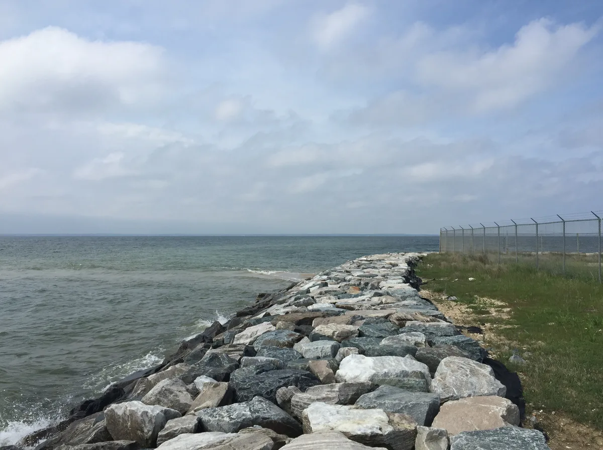 A long riprap jetty leads out into gray-green Chesapeake water beneath a broad, cloud-filled sky, with a chain-link fence and grassy strip running along the right edge. The flat horizon and open water create a windswept, expansive coastal scene.