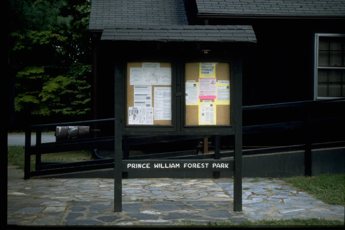 A dark wooden park bulletin board stands in front of a rustic cabin, with maps and notices posted behind glass and “PRINCE WILLIAM FOREST PARK” painted across the base. Stone paving and a black railing frame the foreground, with leafy trees and the cabin’s shingled roof in the background.