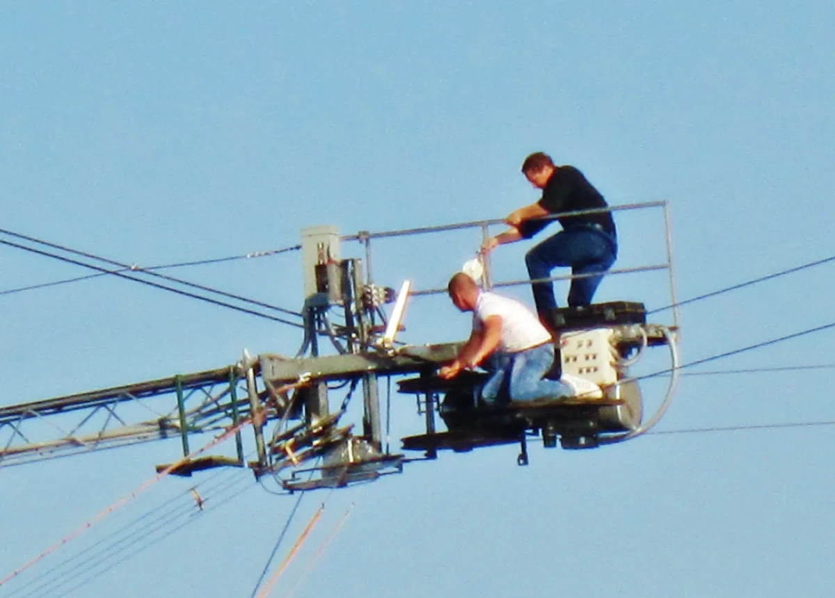 Two workers kneel on a suspended lift high above the ground, repairing a cable against a clear blue sky. The metal boom, tangled wires, and harnessed platform create a stark industrial scene with no landscape visible.