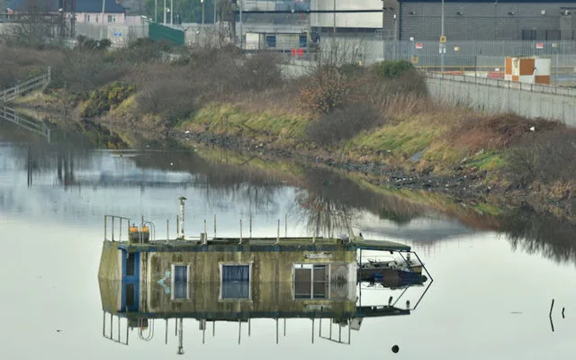 A small houseboat floats on still water, its weathered roof and blue-trimmed doors reflected almost perfectly in the calm surface. Brown winter reeds and grassy banks line the canal, with industrial buildings and fencing in the background.