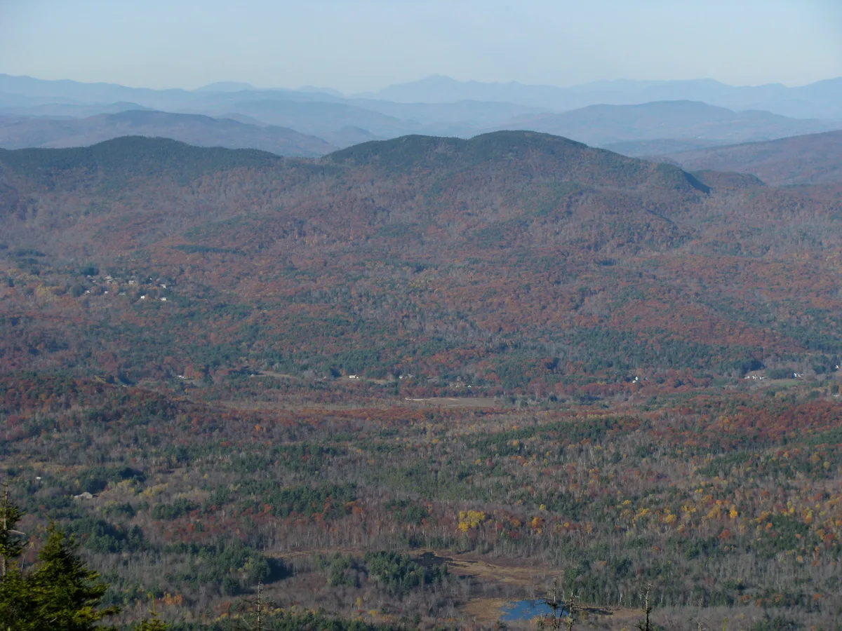 Wide morning view over a broad, forested valley with layered Blue Ridge ridges fading into a pale blue horizon. The slopes are patchworked with autumn color, and a small reservoir glints near the bottom edge beneath the hills.