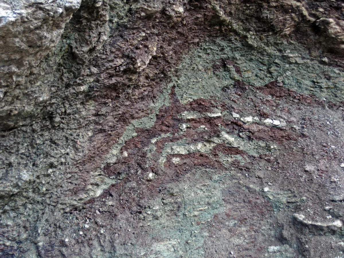 Close-up of a rough, weathered rock face with mottled gray, green, and rust-red mineral stains, layered with cracked textures and scattered pale flecks. The tight framing emphasizes the surface patterns and erosion rather than any distant view.
