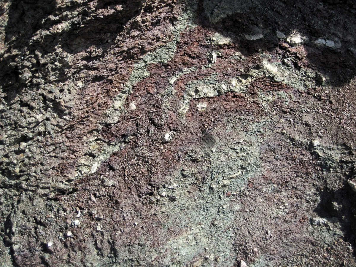 Close-up of a rugged rock surface with mottled reddish-brown and gray-green patches, rough pits, and scattered pale mineral flecks catching the light. The textured stone fills the frame, with shadowed edges emphasizing its uneven, weathered surface.