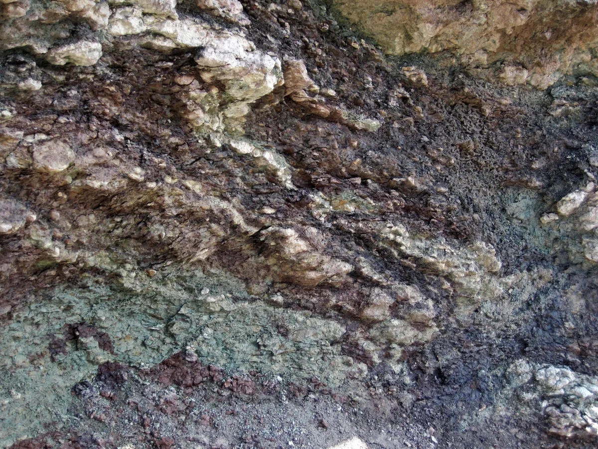 Close-up of a rough rock outcrop with layered gray, brown, and rust-colored textures, mottled with pale mineral veins and weathered pits. The surface fills the frame, creating an abstract geological pattern instead of a wider landscape view.