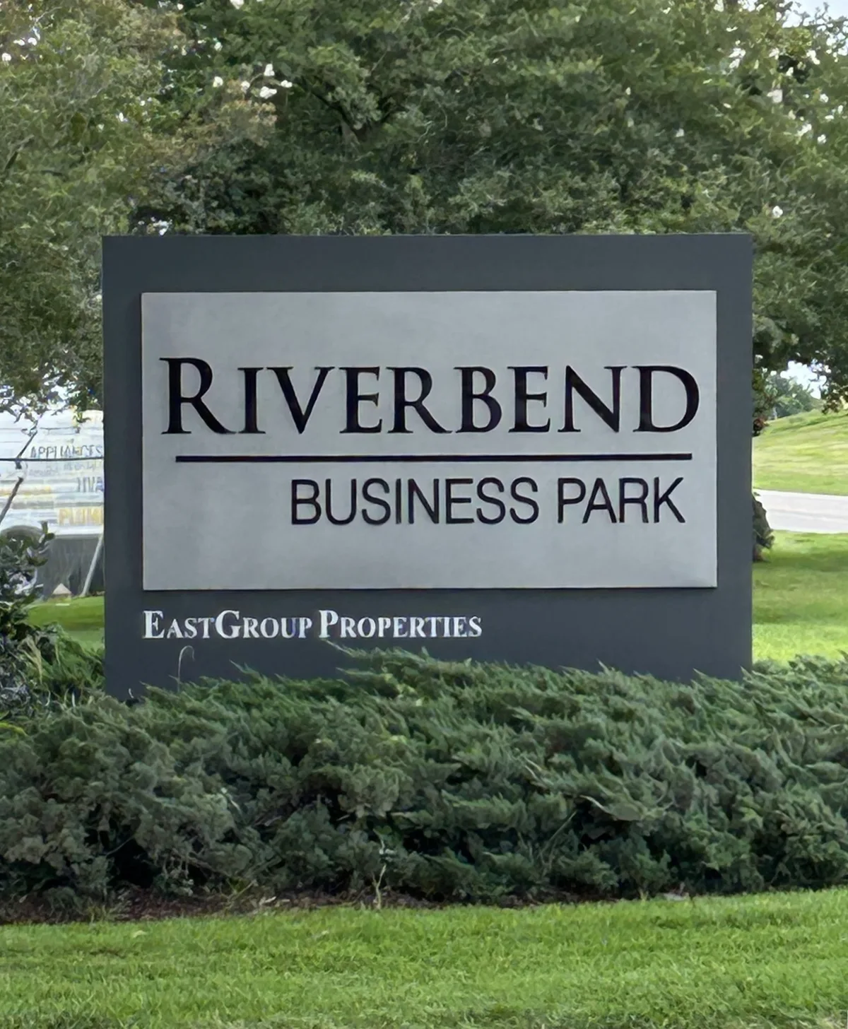 A large roadside sign reading “RIVERBEND BUSINESS PARK” stands in front of trimmed shrubs and bright green grass, with leafy trees filling the background. The scene is evenly lit and looks more like a commercial entrance than a park trail or river view.