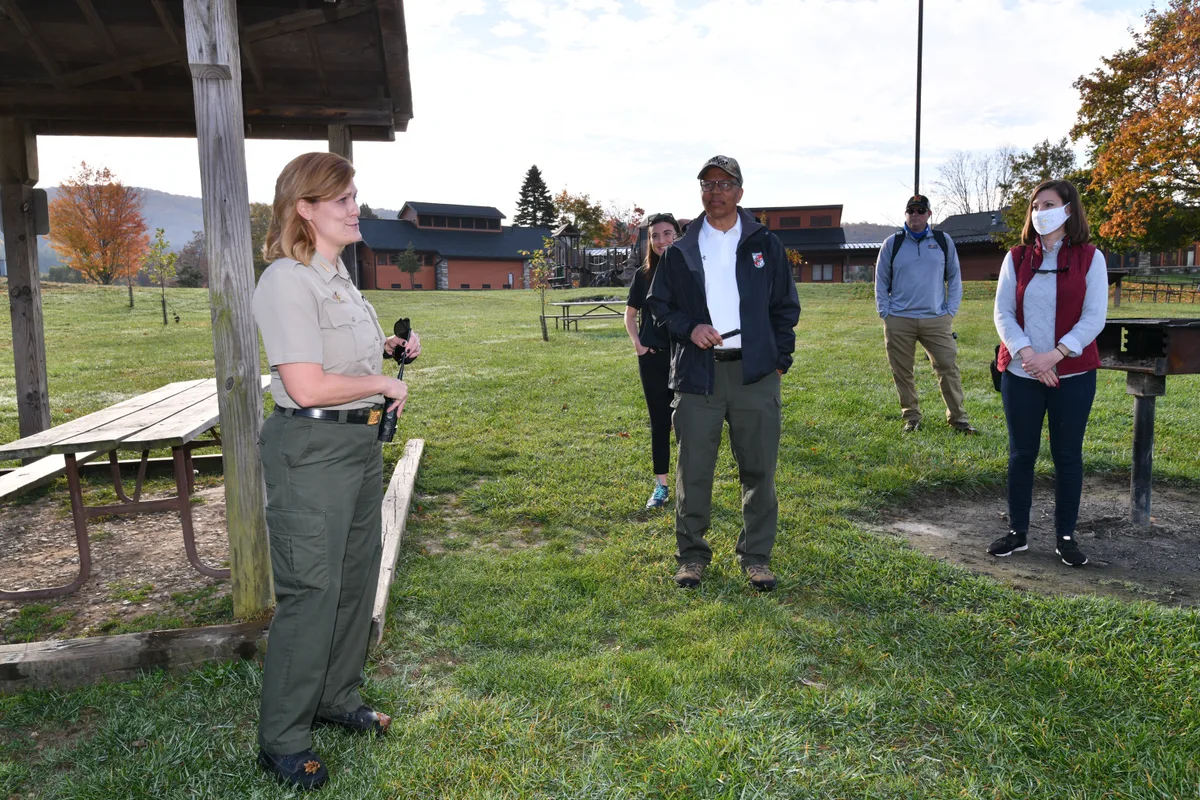 A park ranger in uniform speaks to a small group standing on a grassy lawn beside a picnic shelter and grill, with low buildings and autumn trees in the background. Soft morning light and a pale sky give the scene a calm, open feel.