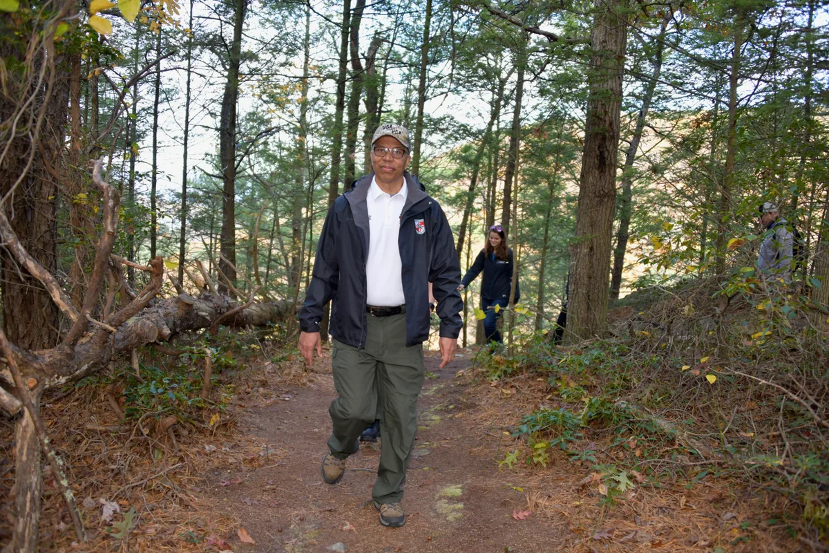 A man walks toward the camera on a wooded trail, framed by tall pines and dense undergrowth. Soft morning light filters through the trees, with a glimpse of the lake and distant shoreline visible between the trunks.