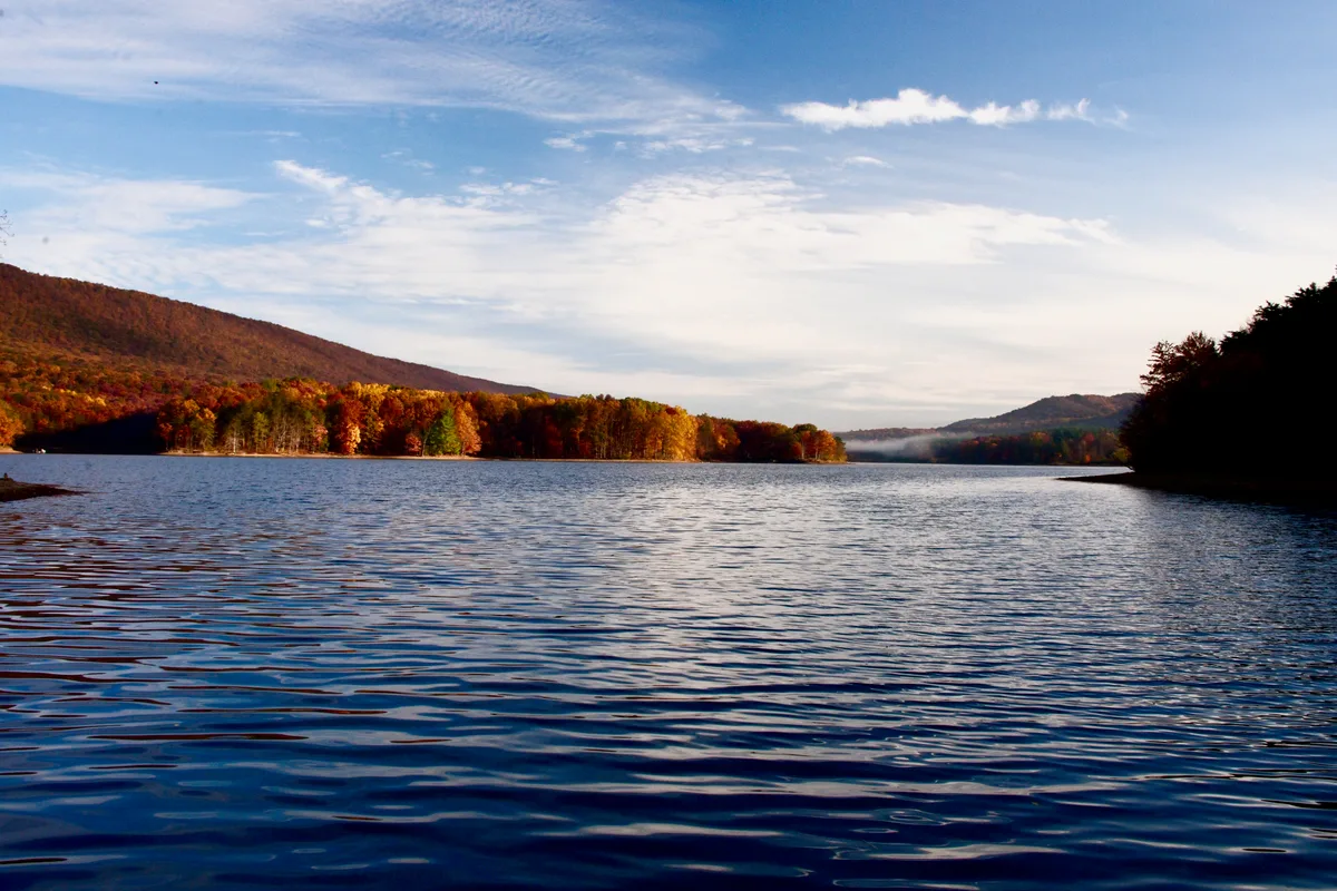Calm blue Lake Habeeb fills the foreground, with gentle ripples reflecting a bright morning sky. Forested ridges in early autumn color frame the narrow mountain gap, with a darker wooded shoreline on the right and a low misty haze in the distance.