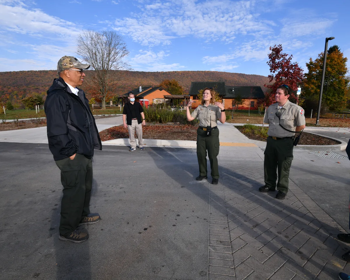 Park staff in uniform stand in a paved lot near Rocky Gap’s visitor buildings, talking under a bright blue morning sky. Fall-colored hills rise behind them, with a few bare trees and autumn leaves framing the scene.
