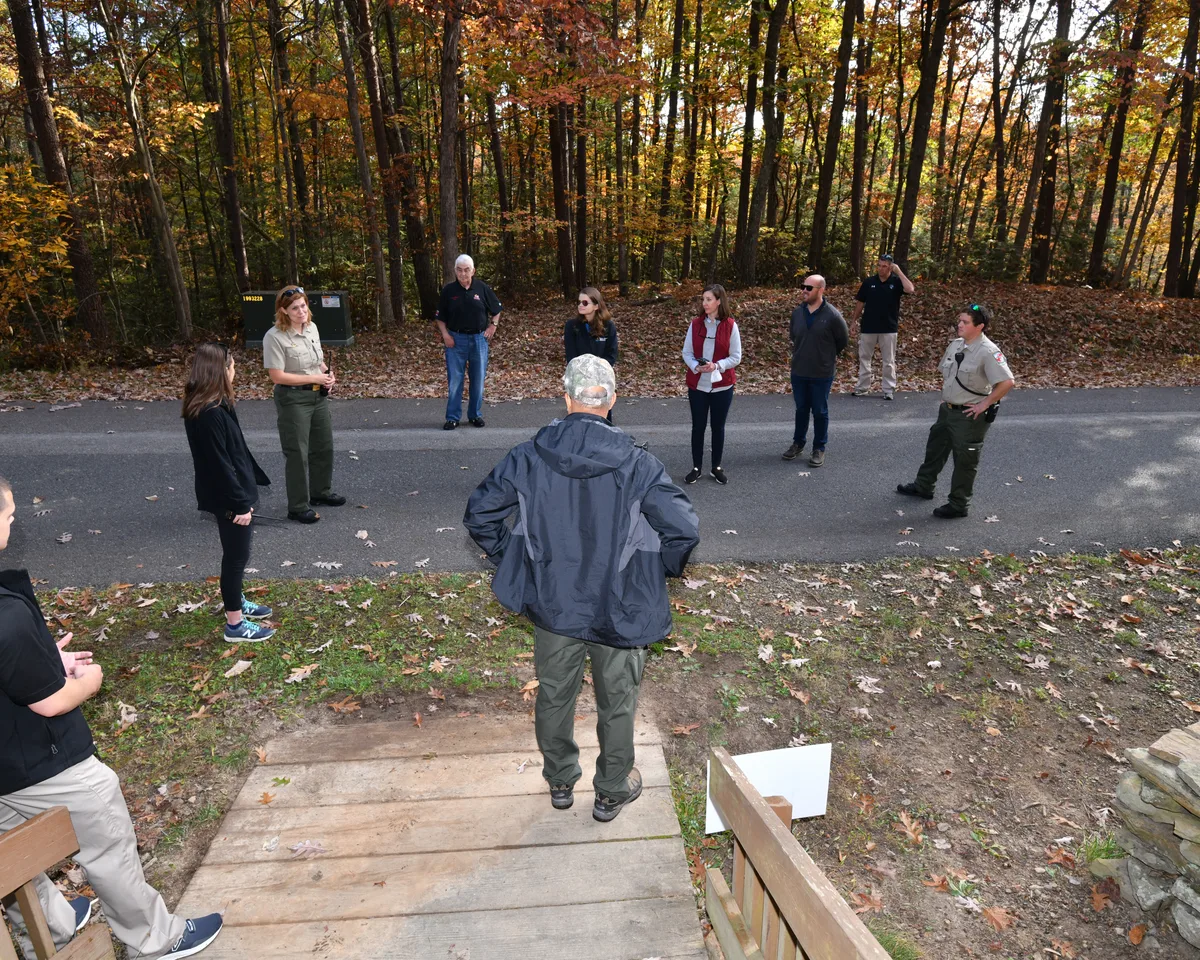 A small group stands in a wooded parking area at Rocky Gap State Park, gathered on a paved road under tall autumn trees with orange and yellow leaves. Two uniformed park staff members appear to be leading the discussion while others listen in a loose circle.