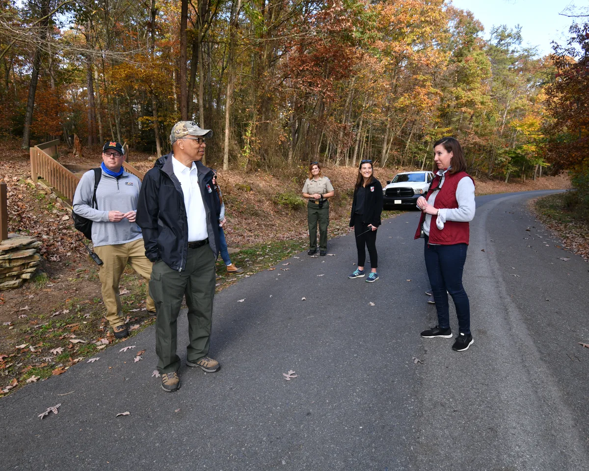 Several people stand talking on a paved park road at Rocky Gap State Park, surrounded by autumn woods with orange and gold leaves. A wooden railing, parked white vehicle, and leaf-strewn ground frame the scene under soft morning light.