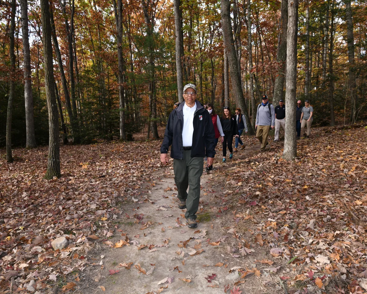 A group of hikers walks along a narrow dirt trail through a wooded forest blanketed in fallen autumn leaves, with tall gray trunks and orange-brown foliage filling the background. The lead man in a dark jacket and cap stands out in the foreground as the others follow behind in soft morning light.