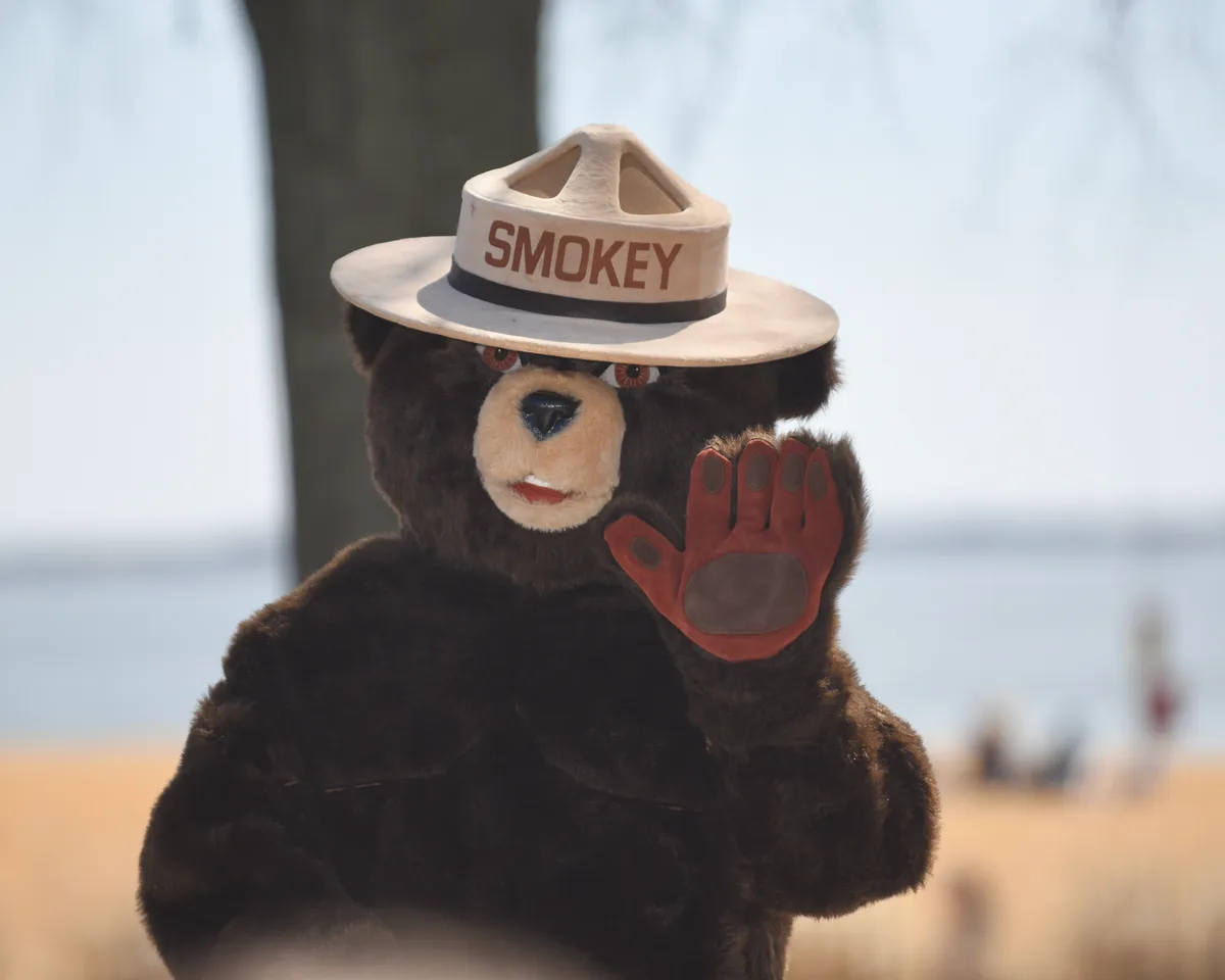 A Smokey Bear statue wearing a ranger hat and raised paw stands in the foreground, with the Chesapeake Bay softly blurred behind it. Warm golden light gives the sandy beach and distant water a hazy, late-day glow.