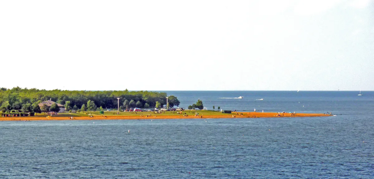 A broad sandy spit at Sandy Point stretches into calm Chesapeake Bay, with tiny beachgoers, a few parked cars, and a tree-lined shoreline under bright golden light. The Bay Bridge itself isn’t visible here, but the open water and low peninsula create a wide waterfront vista.