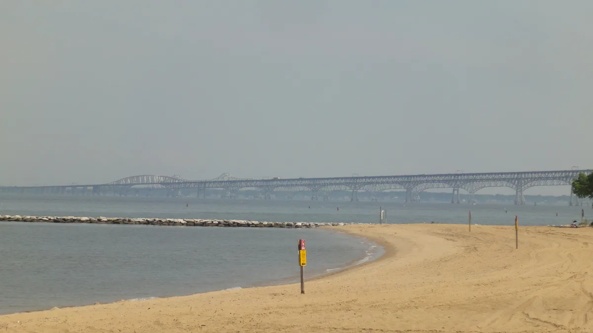 A wide sandy beach curves along the calm Chesapeake Bay, with a low rock breakwater and a few warning posts in the foreground. The Bay Bridge stretches across the hazy horizon under soft, muted golden-hour light.