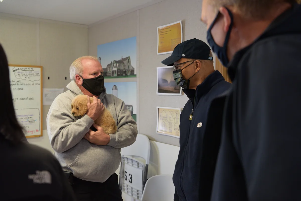 Several masked adults stand in a small indoor room, where a man in a gray hoodie gently cradles a tiny tan puppy against his chest. Framed site photos and notes hang on the beige walls behind them, with white folding chairs stacked along the back.