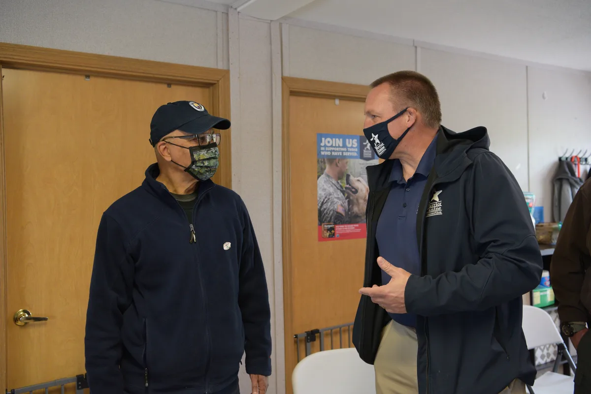 Two masked men stand talking inside a plain meeting room with wooden doors and folded chairs, one gesturing while facing the other. A poster on the back door adds a community-service note to the otherwise simple indoor scene.