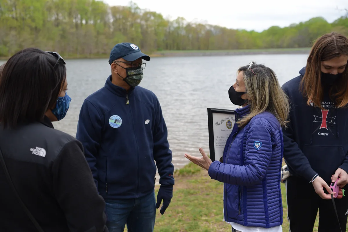 People wearing masks stand on a grassy bank beside the calm reservoir, with wooded spring trees and a pale overcast sky reflected in the water. A woman in a blue jacket speaks with a park staff member while others listen nearby.