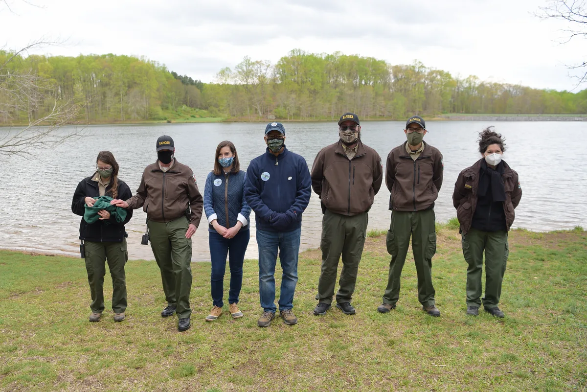 Seven masked park staff and visitors stand in a line on a grassy bank beside the calm reservoir, with spring-green trees and a cloudy sky reflected in the water. The composition feels open and quiet, with the shoreline curving into the wooded background.