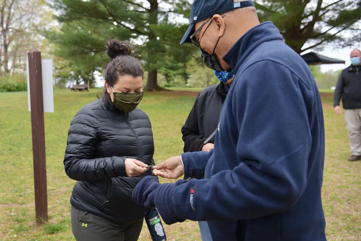 Masked park visitors stand on a grassy clearing beneath tall trees while one person hands a small item to another. Soft morning light and a few scattered signs and picnic shelters suggest a quiet gathering in the park.