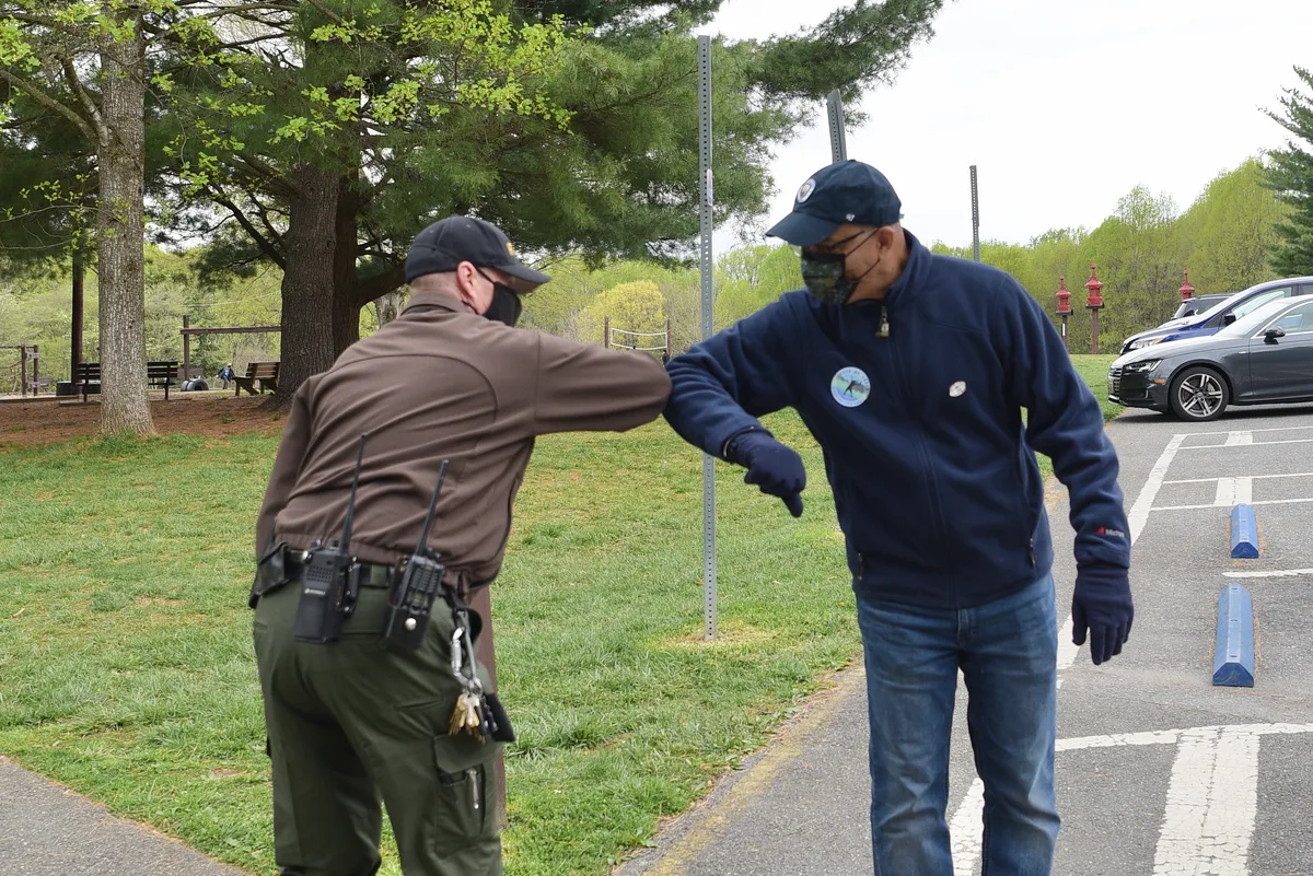 Two masked men on a park path greet each other with an elbow bump beside a grassy lot and tall pines. Parked cars, picnic tables, and a line of spring-green trees frame the scene under an overcast morning sky.