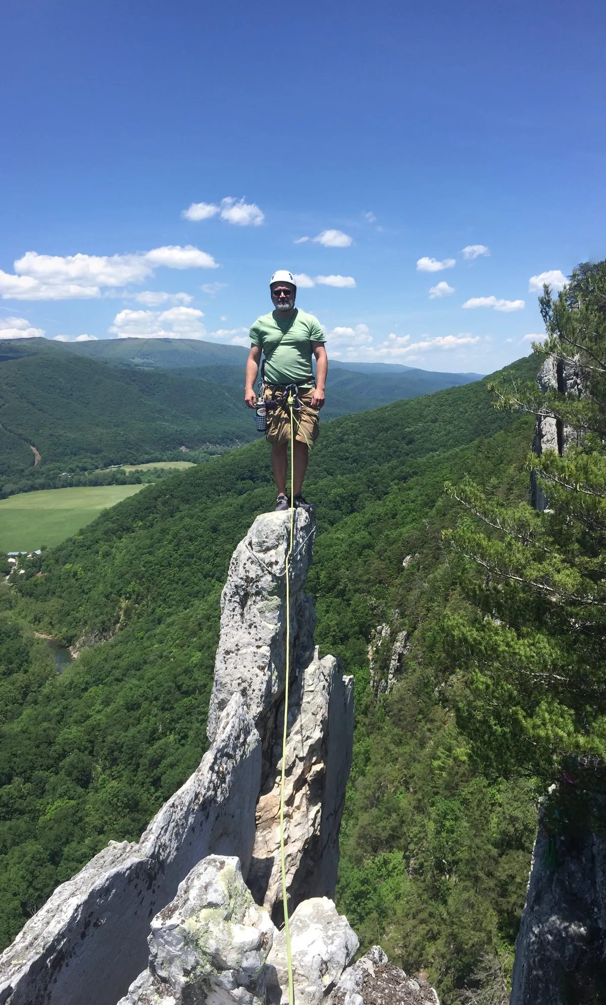 A climber in a helmet stands on the narrow quartzite tip of Seneca Rocks, with a rope hanging straight down the fin. Green mountain ridges and a bright blue sky with scattered clouds stretch behind the dramatic summit.