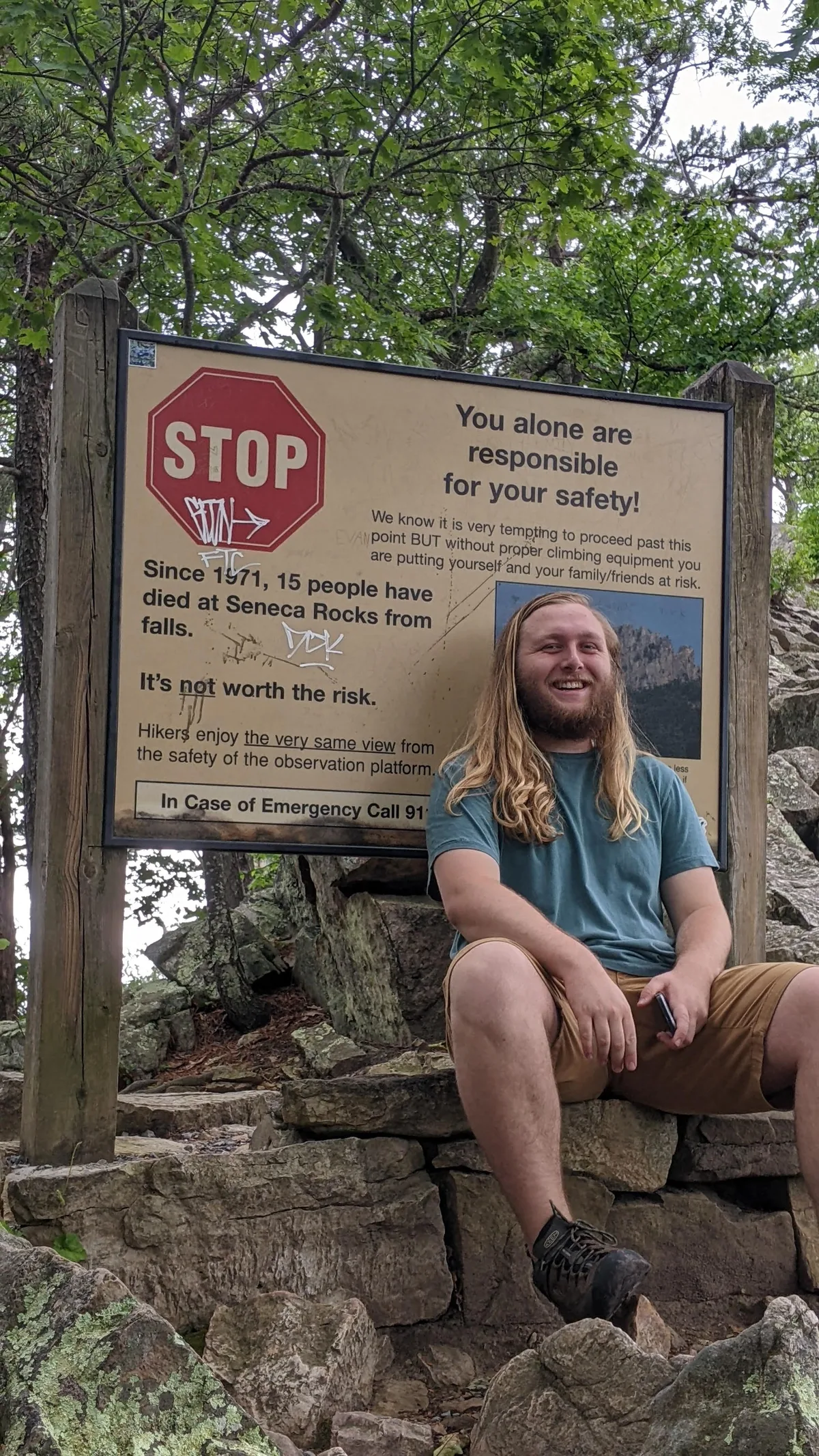 A smiling man sits on stacked stone steps beside a large warning sign at Seneca Rocks, with leafy trees and rough quartzite boulders filling the shaded background. The sign’s bold red STOP symbol and safety message stand out against the natural setting.