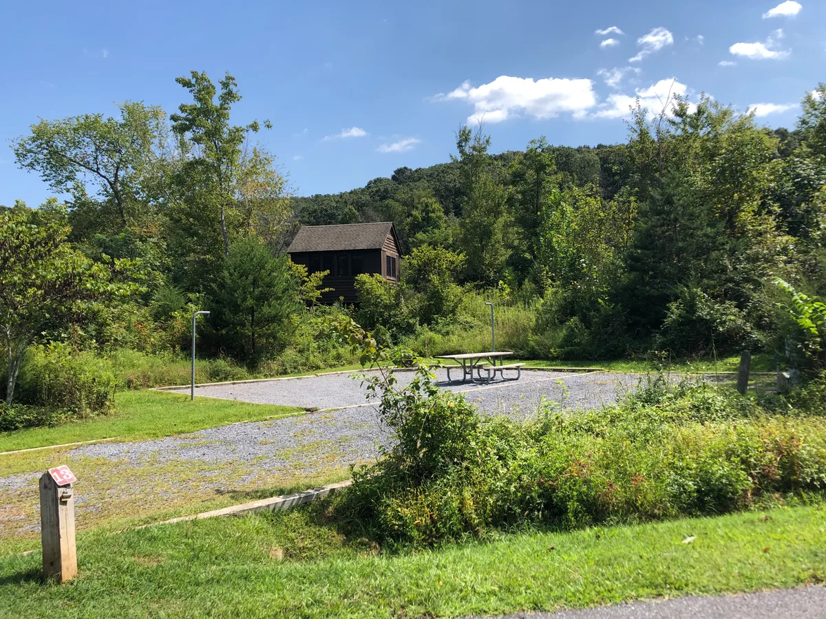 Gravel campsite 13 sits in a sunny clearing surrounded by dense green trees, with a picnic table and water spigots beside a small brown cabin tucked into the hillside. A blue morning sky with a few white clouds hangs over the wooded ridgeline in the background.