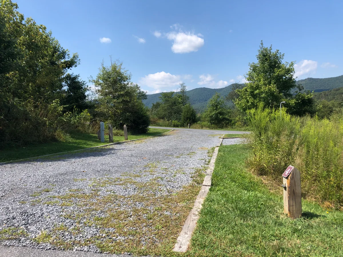 A gravel campsite pad sits in bright morning sun, bordered by mown grass, tall weeds, and leafy trees, with blue mountains rising across the background under a few small clouds. Wooden post markers and a narrow paved edge frame the open site, giving it a quiet, rural feel.