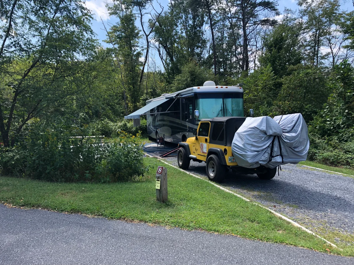 A yellow Jeep and a large motorhome are parked at a shaded campsite, with the Jeep’s rear cargo covered in a gray tarp. Dense green trees and shrubs surround the gravel pad, and a site marker labeled 8 sits beside the grass.