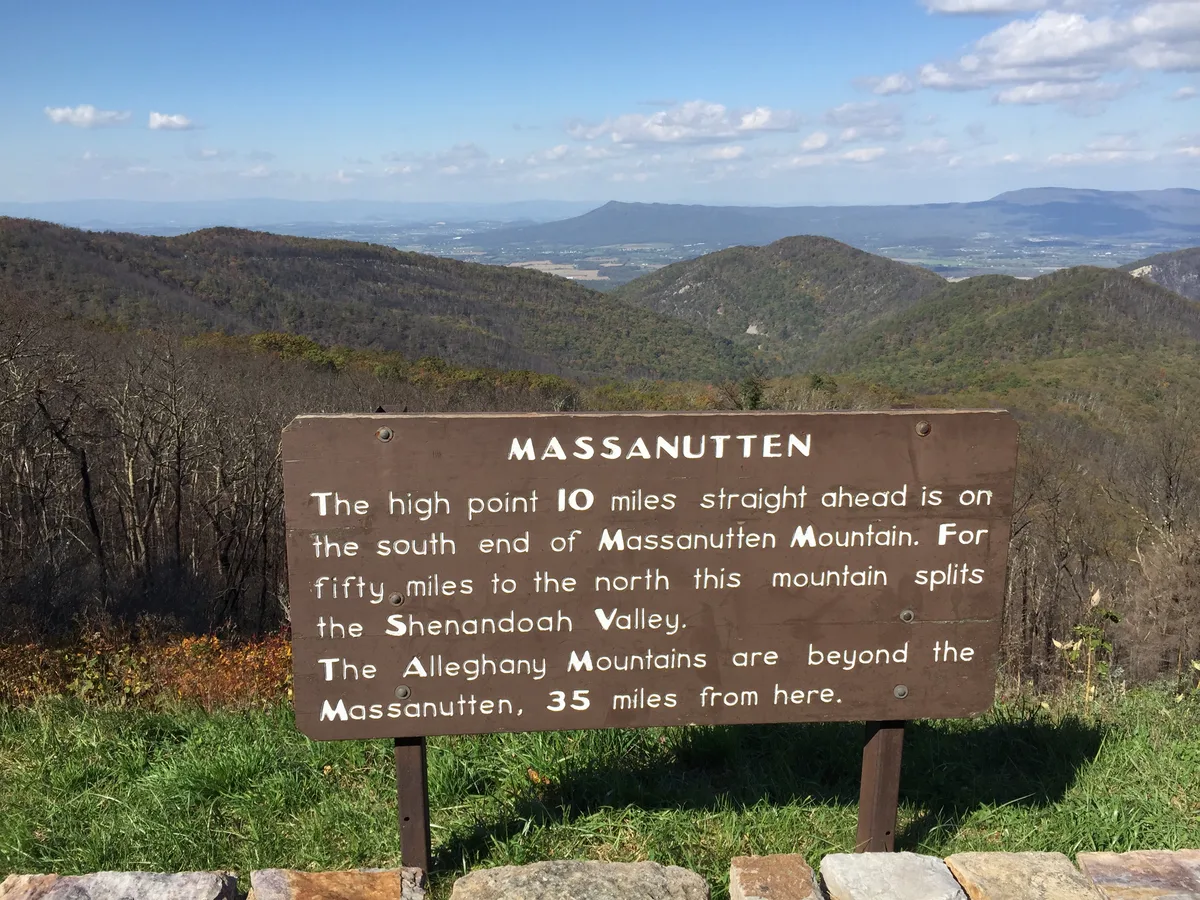 A wooden Massanutten overlook sign stands in the foreground above a grassy roadside pull-off, with layered blue-gray ridges and the wide Shenandoah Valley stretching into the distance under a bright, partly cloudy sky. Bare late-autumn trees and muted green hills frame the view, emphasizing the mountain’s broad sweep across the valley.