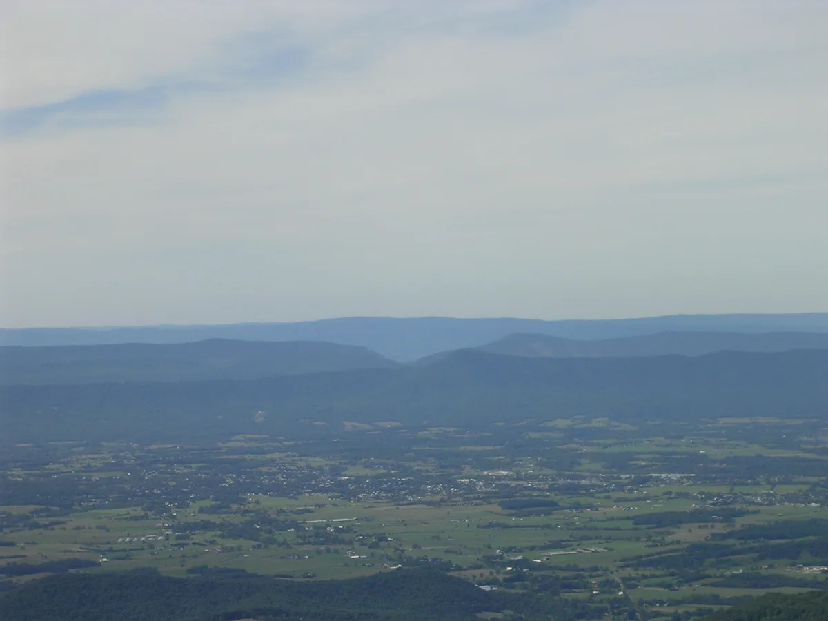 A wide hazy overlook across the Shenandoah Valley shows layered blue ridgelines in the distance and a patchwork of green farmland and small towns below. The scene is soft and atmospheric under pale sky light, with the valley stretching out in broad horizontal bands.