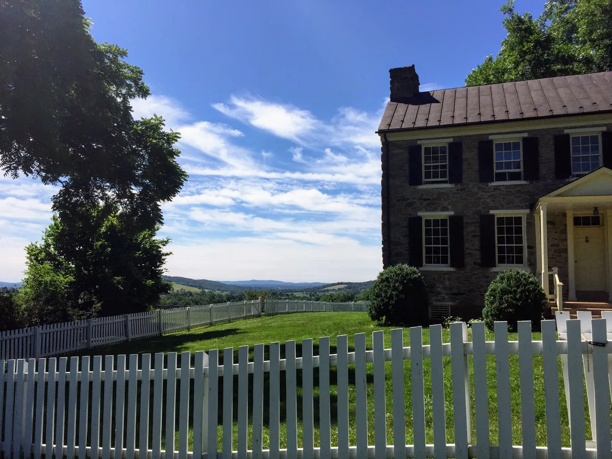 A stone historic house with dark shutters sits behind a white picket fence on a sunny ridge, with a large tree framing the left side and green lawns falling away toward distant Blue Ridge hills. Bright blue sky and wispy clouds open over the sweeping valley view.