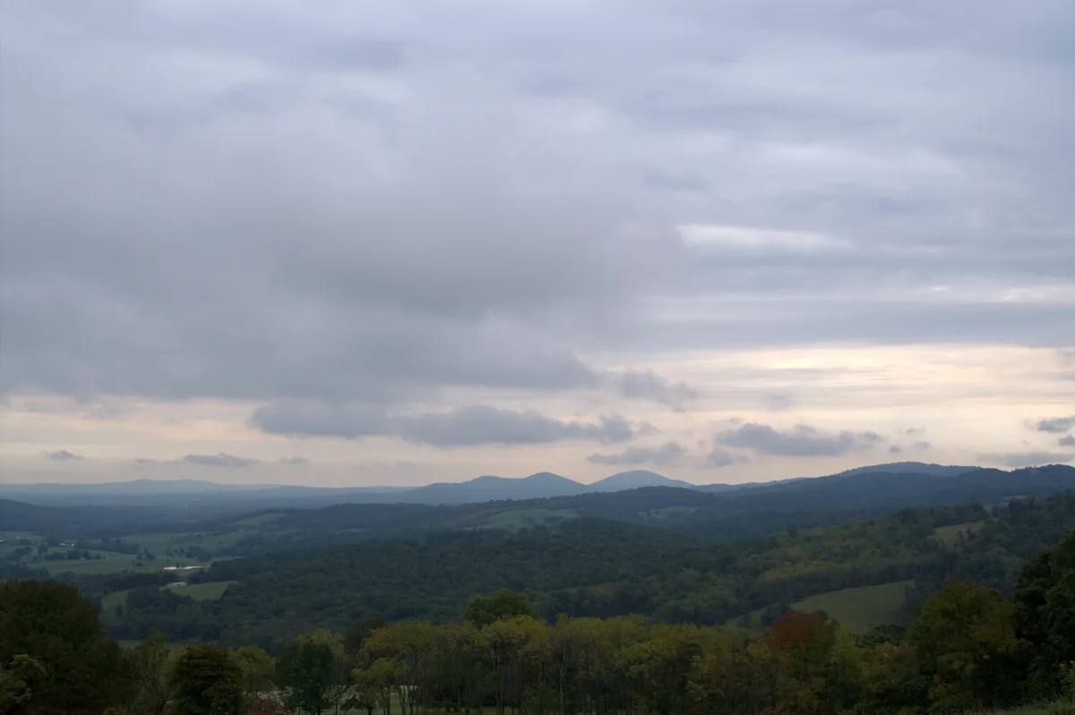 Soft evening light falls over layered Blue Ridge hills and a wide green valley, with dark forested ridges in the foreground and low, gray clouds stretching across the sky. The distant horizon glows faintly near sunset, giving the overlook a calm, pastoral feel.