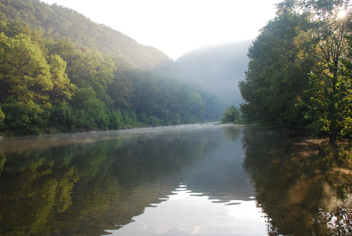 A calm river runs through a narrow, forested canyon, with mist softening the distant ridge and morning light glowing through the trees. The still water mirrors the green slopes and bright sky, emphasizing the quiet, remote feel of Smoke Hole Canyon.