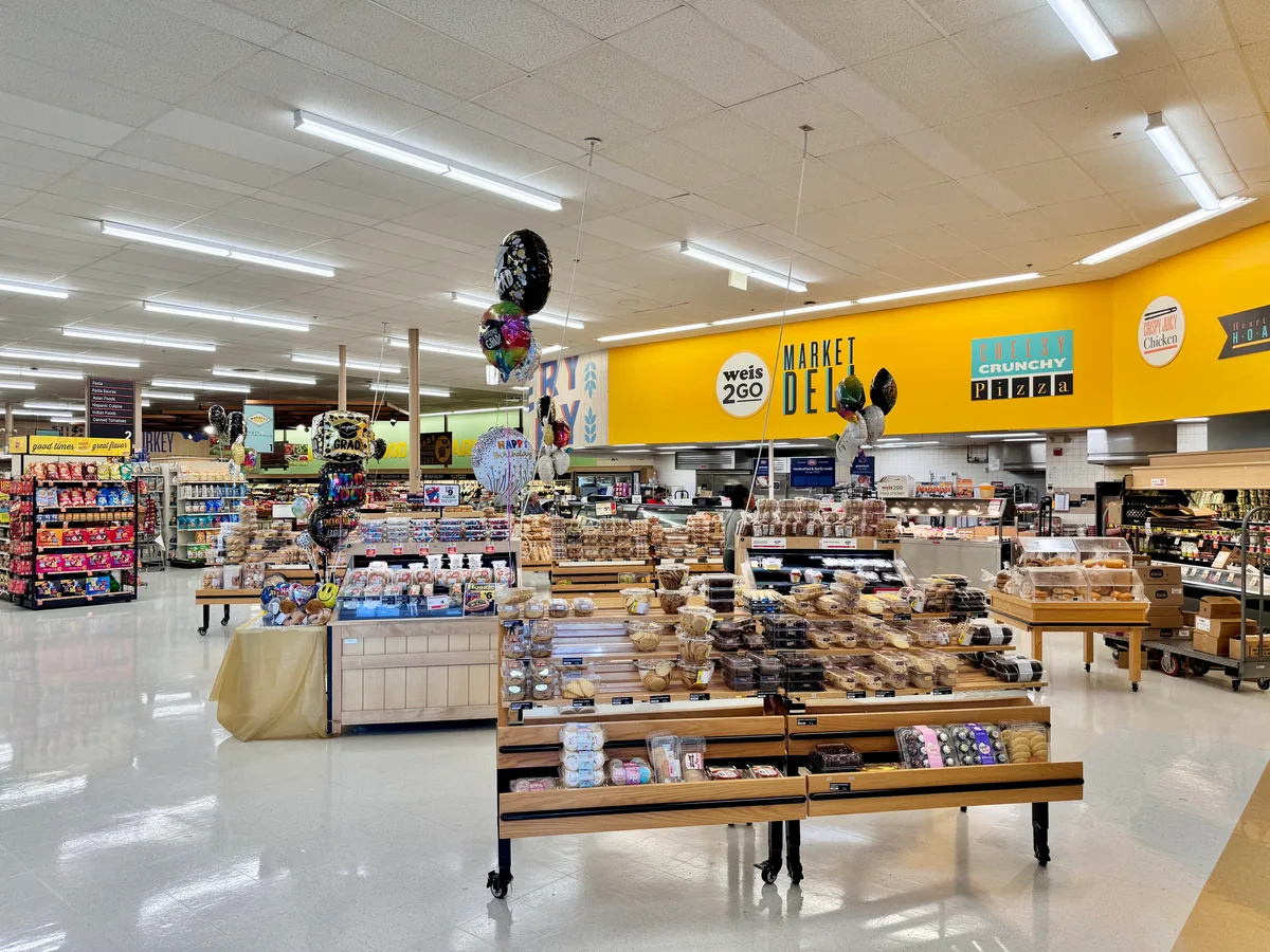 Brightly lit grocery store interior with long fluorescent ceiling lights, a glossy white floor, and a bold yellow wall behind the deli and bakery counters. Balloon clusters and crowded display tables of packaged pastries add a festive, colorful feel.