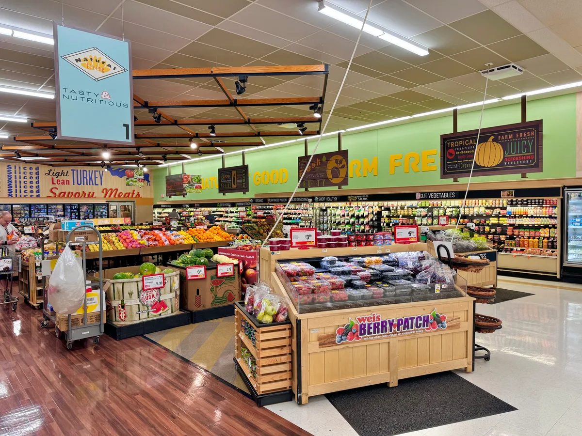 Bright supermarket produce section with wooden display tables piled with berries, melons, oranges, and packaged fruit under large “Nature’s Tasty & Nutritious” and “Farm Fresh” signs. Fluorescent ceiling lights and a glossy wood-look floor give the aisle a clean, colorful, indoor market feel.