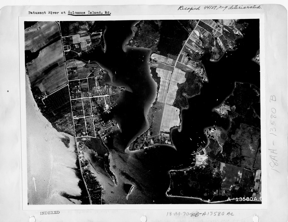 Black-and-white aerial view of Solomons Island and the Patuxent River, showing narrow peninsulas, patchwork fields, roads, and clusters of waterfront homes along the dark channels. The open bay fills most of the frame, with small boats visible as bright specks against the water.