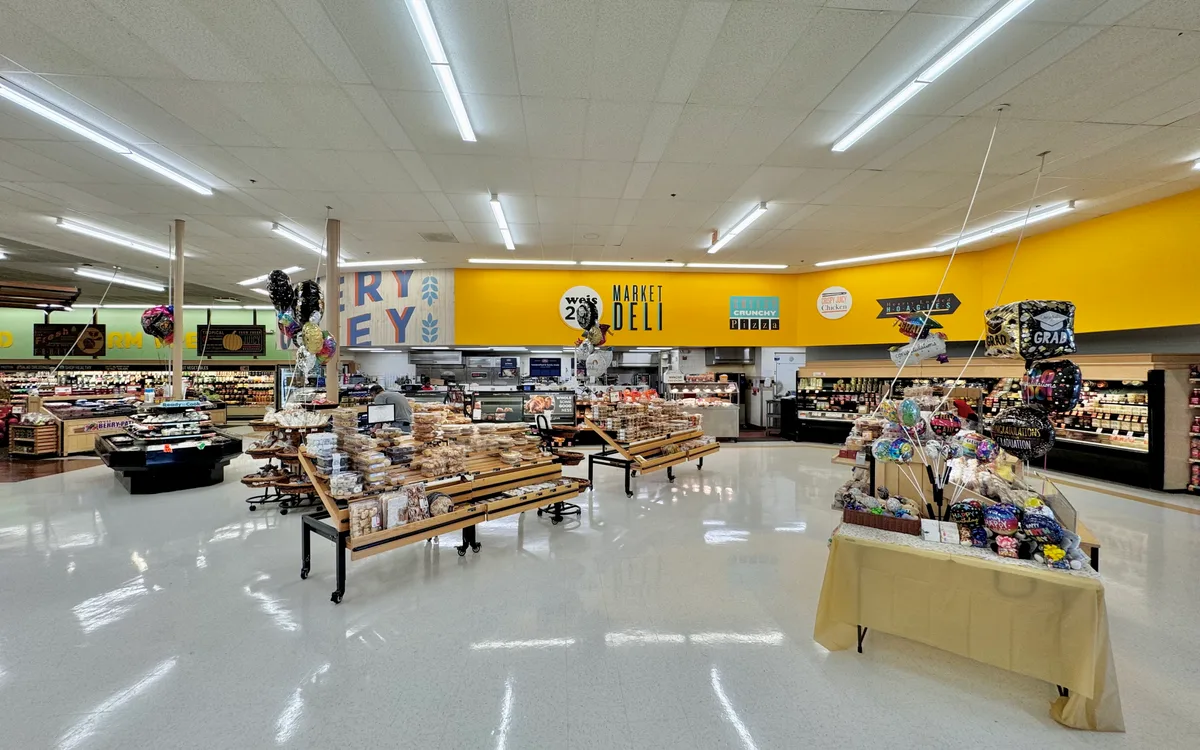 Brightly lit supermarket interior with glossy white floors, yellow accent walls, and rows of bakery and deli displays topped with balloons and gift baskets. The wide view centers on the Weis Market Deli counter under fluorescent ceiling lights, giving the scene a clean, spacious look.