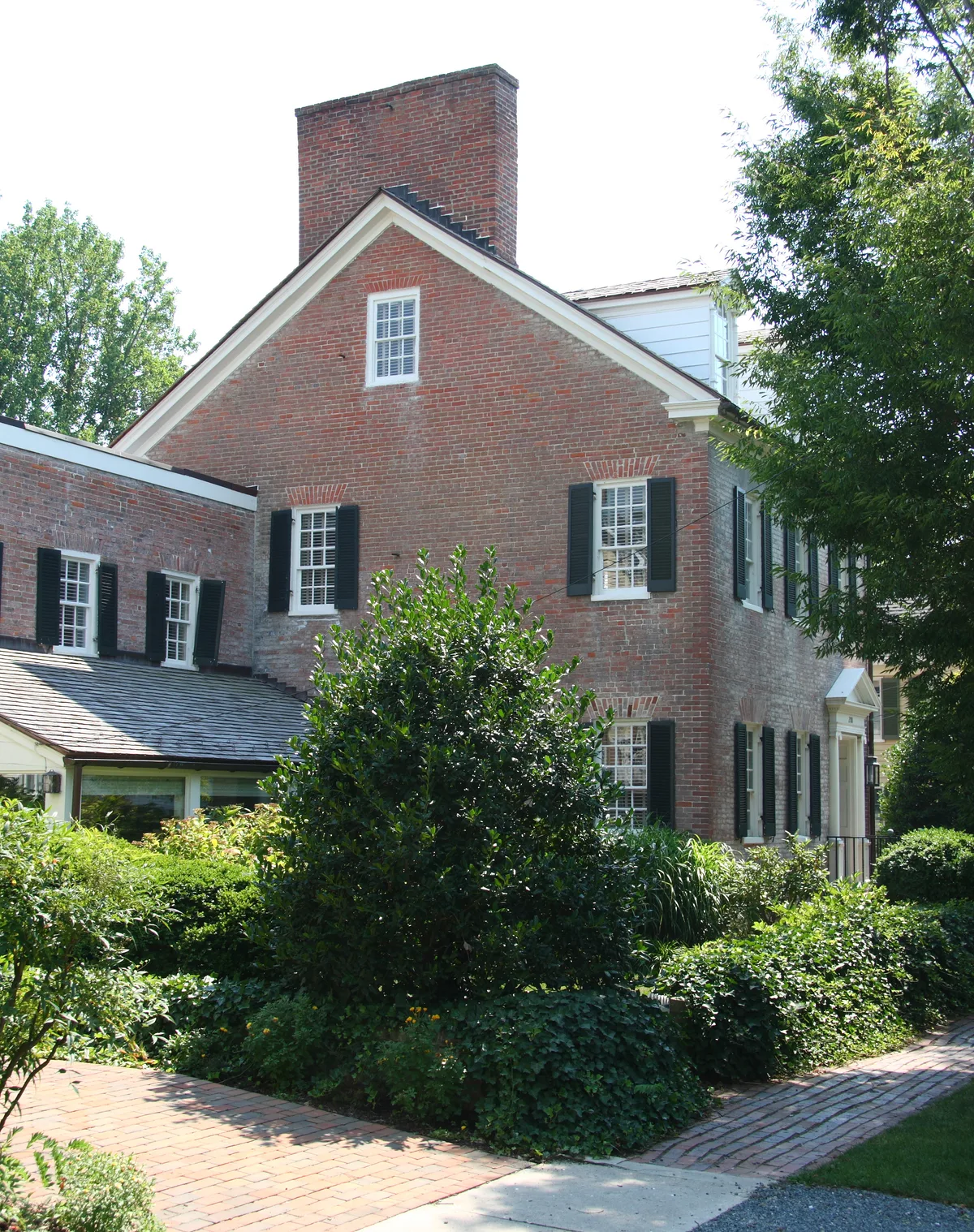 Historic brick Cannonball House rises behind a manicured garden, with black shutters, white trim, and a tall chimney catching bright midday light. Brick walkways and dense green shrubs frame the colonial-style façade in a quiet, well-kept waterfront setting.