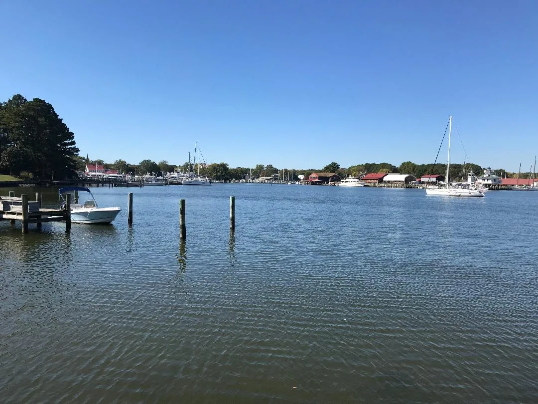 Wide view across calm blue water in St. Michaels Harbor, with a small motorboat tied to a dock at left and a white sailboat floating near the waterfront on the right. Low red-roofed buildings and a line of masts sit along the far shore under a clear blue sky.